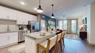 This thoughtfully designed single-floor residence showcases a seamless open-concept layout that connects the kitchen, dining, and living areas into one cohesive, light-filled space. The kitchen is a standout feature, boasting white shaker cabinetry, granite countertops, a subway tile backsplash, stainless steel appliances including a French door refrigerator and range with microwave, and a large central island with pendant lighting and bar seating — ideal for casual dining or entertaining. The living room is anchored by a gallery wall of framed artwork against a rich gray accent wall, with large windows and a glass door providing ample natural light and outdoor access. Wood-look flooring flows throughout the main living areas, adding warmth and continuity. The primary bedroom is a serene retreat featuring generous windows, a ceiling fan, and an en-suite bathroom with granite vanity countertops, a walk-in closet, and a separate water closet. The second bedroom is equally well-appointed with large windows and comfortable proportions. A secondary full bathroom with a tub-shower combination completes the layout. Throughout the residence, consistent use of neutral tones, quality finishes, and carefully considered design details create a polished and inviting atmosphere.