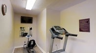 This single-story fitness room offers a practical and functional space designed for cardiovascular exercise and general workouts. The room features light yellow walls with a textured ceiling and is illuminated by overhead fluorescent lighting, providing consistent brightness throughout. A ProForm treadmill takes center stage in the space, complemented by a stationary bike, and an elliptical machine visible in the mirrored wall section. The wall-mounted flat-screen television adds an entertainment element, making workouts more engaging. A large mirror along one wall visually expands the room while allowing users to monitor their form during exercise. The carpeted flooring provides a degree of comfort underfoot. The layout is efficiently organized to maximize the available floor space, accommodating multiple pieces of equipment without feeling overly cramped. Overall, the fitness room presents a straightforward, no-frills environment equipped with essential cardio machines, suitable for residents or occupants seeking convenient access to exercise facilities without leaving the property.