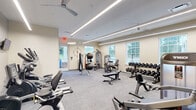 Located at 1000 Cowpen Ct in Williamsburg, Virginia, this fitness center offers a thoughtfully designed exercise space ideal for a well-rounded workout routine. The room features neutral-toned walls, low-profile ceilings with sleek linear LED lighting, and ceiling fans that contribute to a bright, comfortable atmosphere. Large windows along the walls allow natural light to fill the space, creating an open and inviting environment. The flooring throughout is durable, textured carpet well-suited for a fitness setting. The facility is comprehensively equipped for both cardio and strength training. Cardio offerings include treadmills, elliptical machines, and stationary cycling equipment, several of which are positioned near wall-mounted televisions for an engaging workout experience. The strength training area features a multi-function cable machine, a full rack of dumbbells in varying weights, adjustable benches, a leg press machine, and stability balls with resistance accessories. An organized storage rack with labeled bins keeps supplies neatly accessible. Fitness center rules are posted prominently near the entrance, and the space is well-maintained throughout. The layout efficiently maximizes the available floor area, providing ample room to move between stations while accommodating a variety of fitness activities.