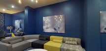 Located on E 22nd St in Bayonne, NJ, this single-floor lounge space delivers a cohesive and visually striking interior defined by deep navy blue walls that set a bold, immersive tone throughout. The space is organized into several distinct functional zones, each thoughtfully furnished to serve different purposes while maintaining a unified aesthetic.

A comfortable lounge area anchors the main space, featuring a modular sectional seating arrangement composed of gray, black, yellow, and patterned upholstered pieces arranged around a round white coffee table. Geometric starburst-patterned area rugs in blue and white ground each seating zone and add an additional layer of visual texture. Large-scale photographic art prints depicting dramatic cloudscapes are displayed prominently across the walls, reinforcing the space's sky-inspired design theme.

A dedicated workspace area includes a mid-century modern wood desk paired with a vibrant yellow molded chair, providing a productive setting within the lounge. Adjacent to the workspace, a wall-mounted flat-screen television sits atop a decorative carved media console, adding an entertainment component to the space.

A separate dining nook features a round walnut-toned table with sleek black and gold-accented chairs, ideal for small gatherings. Recessed lighting throughout provides warm, focused illumination, while hardwood flooring ties the entire space together with warmth and continuity.