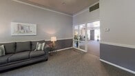This single-floor community space is thoughtfully designed to provide a comfortable and functional gathering environment. The layout is divided into distinct zones, each serving a specific purpose while maintaining a cohesive aesthetic throughout.

The lounge area features plush, dark gray upholstered seating arranged alongside accent tables with warm lamp lighting, creating an inviting atmosphere for relaxation or casual conversation. Framed artwork decorates the textured walls, and large glass-paned doors connect this area to adjacent spaces, allowing natural light to flow through while maintaining visual openness.

The dining area centers around a sturdy wooden high-top table surrounded by cross-back chairs, well-suited for small group gatherings or meetings. A large decorative wall clock serves as a focal point, complementing the neutral two-tone wall treatment featuring a darker wainscoting below and lighter tones above. A window provides natural light and outdoor views, while a water cooler adds everyday convenience.

The kitchenette includes dark cabinetry, a double-basin stainless steel sink, light countertops, and a dishwasher, offering practical utility for events or daily use. Consistent crown molding, carpeted flooring in the common areas, and hardwood-style flooring in the kitchenette tie the space together with a polished, well-maintained finish.
