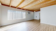 Located in the Canoga Park neighborhood of Los Angeles, this 1-bedroom, 1-bathroom unit offers a distinctive architectural character defined by its signature exposed wood beam ceilings with natural pine tongue-and-groove paneling throughout. The open-concept living and dining area is bathed in natural light through large windows dressed with vertical blinds, and features light-toned luxury vinyl plank flooring that complements the warm wood overhead. A ceiling fan adds comfort to the airy living space, which flows seamlessly toward the entryway and hallway. The galley-style kitchen is well-appointed with rich dark mahogany cabinetry, light stone countertops, a stainless steel sink, and black appliances including a range and refrigerator, all beneath the same signature beamed ceiling. The bedroom offers plush wall-to-wall carpeting, a sliding-door closet, a window with natural light, and its own wall A/C unit for personalized climate control. The full bathroom features tile flooring, a dark wood vanity with a stone countertop, and a tub-shower combination with a sliding glass enclosure. This thoughtfully designed unit delivers a warm, inviting ambiance with consistent architectural details throughout.