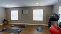 This single-floor yoga and fitness studio offers a calm, focused environment designed to support a variety of movement practices. The open studio space features warm hardwood flooring that provides both comfort and durability, creating a welcoming surface for yoga mats, stretching, and balance training. Neutral taupe-toned walls contribute to a serene, distraction-free atmosphere that encourages concentration and relaxation. Multiple windows fitted with white horizontal blinds line the walls, allowing natural light to filter into the space while offering privacy control as needed. A recessed ceiling light provides additional illumination, ensuring the studio remains well-lit throughout the day. The layout is intentionally open and uncluttered, offering ample floor space to accommodate multiple practitioners simultaneously. Fitness equipment such as yoga mats, foam blocks, and a BOSU balance trainer are present, reflecting the studio's versatility for yoga, balance training, and general fitness. A framed instructional or inspirational piece adorns one wall, adding a purposeful, educational touch to the environment. The clean lines, thoughtful design, and practical amenities make this studio a functional and inviting space for both individual and group wellness sessions.