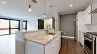Located at 23 North St in Buffalo, NY, this thoughtfully designed single-floor unit blends contemporary finishes with functional living. The open-concept kitchen serves as the heart of the space, showcasing a large island topped with speckled granite countertops, a undermount sink, and sleek white cabinetry. Brushed gold pendant lights add a warm, upscale touch, while stainless steel appliances and a stainless range hood complete the culinary setup. An in-unit washer is conveniently integrated nearby. Rich hardwood flooring flows through the main living areas, complemented by crown molding and recessed lighting throughout. The living area is bright and open, featuring bold black-framed floor-to-ceiling windows that frame striking views of Buffalo's historic neighboring architecture. The bedrooms are finished with plush, multi-toned carpet and black-framed windows that invite natural light. The bathroom is a standout feature, boasting a walk-in glass-enclosed shower clad in dramatic marble-look tile, hexagonal floor tile, and brushed nickel fixtures. The overall aesthetic is clean, modern, and refined, offering a polished urban living experience in the heart of downtown Buffalo.