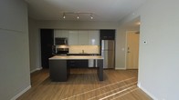 This single-floor space showcases a clean, modern design centered around an open-concept layout. The kitchen serves as the heart of the unit, featuring a striking two-tone cabinetry scheme with dark lower cabinets contrasted by light upper cabinets finished in a smooth, high-gloss style. A central island with granite countertops provides additional workspace and serves as a natural dividing point between the kitchen and the adjoining living area. Stainless steel appliances, including a refrigerator, microwave, and range, lend a polished, professional aesthetic to the space. A neutral tile backsplash runs the length of the counters, adding texture while maintaining a cohesive look. Track lighting installed on the ceiling illuminates the kitchen efficiently, enhancing the contemporary feel. Warm-toned hardwood flooring flows throughout, adding a natural element that softens the otherwise modern palette. The open floor plan creates a seamless connection between the kitchen and living areas, promoting a spacious and airy ambiance. A wood-paneled entry door is visible, completing the well-appointed and thoughtfully designed interior.