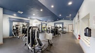 Located at 1700 Old England Loop in Sanford, Florida, this fitness center offers a generously sized, single-floor exercise room designed to support a comprehensive workout experience. The space is immediately distinguished by its bold navy blue vaulted ceiling, which adds visual drama and a sense of height, complemented by recessed lighting that illuminates the entire floor evenly. The light gray walls provide a clean, neutral backdrop that keeps the space feeling open and well-organized.

The exercise room is outfitted with an extensive selection of strength training machines, including multi-station cable units, chest press, leg press, and various resistance machines suited to full-body workouts. Cardio equipment is equally well-represented, with multiple treadmills and stationary bikes positioned throughout the room. Full-length mirrors framed by elegant blue-painted pilasters with decorative crown molding are strategically placed to aid form and enhance the sense of space.

The durable dark rubber flooring provides both safety and practicality underfoot. Natural light filters in through windows, including an arched feature window, adding brightness to the interior. A pass-through window and French door connect the fitness room to an adjacent area, ensuring easy access. The overall ambiance is polished yet functional, making this fitness center a well-appointed facility for residents of the community.