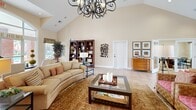 Located at 901 Forest Lake Ct in Newport News, Virginia, this single-floor space offers a well-appointed and thoughtfully designed layout ideal for community gatherings or professional use. The centerpiece of the space is a generously sized living room distinguished by soaring vaulted ceilings and an elegant wrought-iron chandelier that commands attention. Large windows allow natural light to fill the room, highlighting the light tile flooring, warm neutral walls, and comfortable seating arrangements. A built-in entertainment center and decorative bookshelves add both function and character, while area rugs and curated décor create an inviting ambiance. The kitchen and living area are seamlessly connected, with the kitchen showcasing rich cherry wood cabinetry, dark granite countertops, a stainless steel refrigerator, and a peninsula with seating — ideal for casual interaction. A dedicated office provides a more intimate setting, furnished with a round conference table, multiple chairs, and a window overlooking the exterior greenery, making it well-suited for private meetings or consultations. Throughout the space, consistent tile flooring, recessed lighting, and warm beige tones contribute to a cohesive and professional yet welcoming atmosphere.