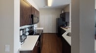 This single-story residence offers a thoughtfully designed layout with distinct living spaces connected by rich hardwood flooring throughout the main areas. The open-concept living room features neutral warm-toned walls, a textured ceiling, and ample natural light, with a defined dining nook accommodating a bistro-style high-top table and bar seating alongside a pass-through opening to the kitchen. The kitchen is outfitted with dark espresso cabinetry, light-colored countertops, stainless steel appliances including an electric range and microwave, and a double-door pantry closet, providing generous storage and counter space in a galley-style configuration. Two bedrooms offer carpeted flooring and notable design details: one features a bold dark accent wall with decorative round mirrors and a closet with double doors, while the other benefits from large windows framing panoramic mountain views, a soft grey accent wall, and a teal feature wall option. Both rooms receive generous natural light. The full bathroom includes a dark wood vanity with an undermount sink, framed mirror with overhead lighting, a full-size soaking tub and shower combination with a curtain rod, and coordinating framed wall art, creating a clean, contemporary aesthetic throughout.