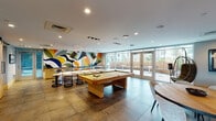 Located at 417 W Peace St in Raleigh, NC, this single-floor amenity lounge is a thoughtfully designed social and recreational space that combines modern aesthetics with a lively, engaging atmosphere. The open-plan layout is anchored by a bold, large-scale abstract mural in vibrant hues of yellow, blue, green, and orange, serving as a striking focal point throughout the space.

The main gathering area features a full-size pool table set atop large-format tile flooring, complemented by a bar-height counter with sleek stools — ideal for socializing. A dedicated gaming nook houses classic arcade cabinets alongside a large wall-mounted television, with comfortable, low-profile lounge chairs and round ottomans creating a relaxed viewing and gaming environment. Built-in shelving and curated artwork add a refined, residential touch.

A cozy semi-private booth, finished in deep forest green with a curved, tufted banquette and a marble-top table beneath a woven pendant light, offers an intimate retreat within the larger space. Floor-to-ceiling glass walls and French doors flood the interior with natural light and open directly onto an outdoor terrace, extending the social space outside. A suspended rattan egg chair near the entrance adds a playful, design-forward accent. The overall ambiance is energetic yet inviting, balancing entertainment and comfort seamlessly.