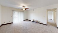 Located at 106 Castle Dr in Dothan, Alabama, this one-bedroom, two-bathroom unit offers a comfortable and functional single-floor layout. The living area features plush wall-to-wall carpeting, a ceiling fan with light fixture, and a sliding glass door framed by vertical blinds that opens to a private balcony, allowing natural light to fill the space. The kitchen is well-equipped with rich, dark wood cabinetry, granite-style countertops, a tile mosaic backsplash, hardwood-style flooring, and black appliances including an electric range and dishwasher — providing ample storage and counter space for everyday cooking. A connecting hallway with hardwood-style flooring leads to the bathrooms, one of which features a shower-tub combination with crisp white surroundings and a vanity with granite-style countertops. The generously sized primary bedroom is carpeted and includes a ceiling fan, a natural light-admitting window, and access to a dedicated walk-in closet alongside an additional louvered-door closet. Throughout the unit, neutral-toned walls, dark baseboards, and textured ceilings contribute to a clean, cohesive aesthetic. The layout flows efficiently through a central hallway connecting all rooms.