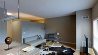 This single-floor space showcases a thoughtfully designed interior with a modern, minimalist aesthetic. The living area is the centerpiece, characterized by smooth white ceilings with subtle recessed lighting that casts a warm glow, contrasting beautifully against the earthy taupe and grey walls. The color palette is cohesive and understated, creating a calm and inviting atmosphere throughout.

The living room is furnished with a sleek, low-profile grey sofa adorned with patterned throw pillows, complemented by a matching grey accent chair that adds a mid-century modern touch. A rectangular glass-top coffee table with a natural wood frame anchors the seating arrangement, while a cowhide area rug layered over a lighter base rug adds texture and visual interest to the hardwood floor beneath.

Ambient lighting is provided by a classic arc floor lamp and a compact table lamp on a round side table, contributing to the warm and relaxed mood of the space. The open layout and thoughtful furniture placement give the room a sense of spaciousness and flow. Overall, this space presents a well-curated, contemporary living environment that balances style and comfort with a clean, uncluttered design sensibility.