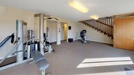 This fitness center offers a practical and well-organized space for residents to maintain an active lifestyle. The room is finished with neutral-toned walls and wall-to-wall carpeting, creating a clean and comfortable environment underfoot throughout the workout area.

The space is outfitted with a variety of exercise equipment, including a multi-station cable weight machine for comprehensive strength training, a stationary recumbent bicycle, and a treadmill positioned near the windows. A dedicated staircase with a warm wood railing is prominently featured, providing access to additional areas of the building and adding an architectural element to the room.

Natural light streams in abundantly through a series of large windows along one wall, offering views of the surrounding exterior grounds and keeping the space feeling bright and open. Flush-mounted ceiling light fixtures supplement the natural light for use during all hours.

Safety features are thoughtfully incorporated, including a clearly marked exit door, emergency exit signage, and a wall-mounted fire extinguisher. A fitness center rules and information board is posted on the wall, ensuring users are informed of facility guidelines. The layout is open and uncluttered, allowing ample room to move freely between equipment and conduct stretching or floor exercises with ease.