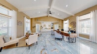 This thoughtfully designed single-floor space makes a striking first impression with its expansive, open layout and soaring vaulted ceilings accented by white exposed beams and a contemporary ceiling fan. Throughout the entire space, large-format blue and gold marble-look porcelain tile flooring creates a cohesive, luxurious foundation that reflects natural light beautifully.

The main area serves a dual purpose as both a welcoming reception and collaborative workspace. A warm gold-toned reception counter lined with white cabinetry anchors one wall, adorned with starburst wall art and pendant lighting. Nearby, a substantial reclaimed wood desk with ergonomic seating accommodates an active workstation, while comfortable lounge seating arrangements invite visitors to relax.

An adjoining lounge area benefits from generous natural light streaming through large windows dressed in white curtains, alongside French doors that open to the outdoors. A cozy bistro table and bookshelf add functional charm to this bright nook.

A compact but fully equipped kitchenette features white shaker cabinetry, quartz countertops, stainless steel appliances, and a peninsula workspace. A utility hallway with additional cabinetry and a sink provides practical support space.

Warm golden-tan walls throughout create an inviting, professional ambiance, complemented by tasteful decorative accents, potted greenery, and abundant recessed lighting.