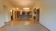 This single-floor space is designed with an open-concept layout that promotes a sense of spaciousness and easy flow between living areas. The kitchen serves as the heart of the home, anchored by a substantial central island that provides both workspace and a natural gathering point. Warm honey-toned wood cabinetry lines the kitchen walls, complemented by black appliances including a built-in microwave and range. Pendant lighting and an elegant chandelier hang overhead, adding warmth and definition to the kitchen and dining area. The vaulted ceiling rises overhead, enhancing the open, airy atmosphere of the main living space. A combination of tile-style flooring in the kitchen and dining zone transitions into plush carpeting in the living area, delineating each functional space while maintaining a cohesive feel. A built-in desk or utility nook with matching cabinetry is tucked to one side, offering a practical workspace. Multiple interior doors lead to additional rooms, suggesting a well-organized floor plan. Neutral wall tones throughout keep the space feeling bright and inviting, making it a versatile and functional single-level home.