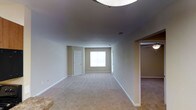 This single-floor residence offers a clean, functional layout designed for comfortable everyday living. The main living area is spacious and open, featuring wall-to-wall carpeting in a warm neutral tone that extends throughout the space, creating a cohesive and inviting atmosphere. The walls are finished in soft, light hues that complement the overall bright and airy feel of the interior.

A centrally positioned window in the main room allows natural light to filter in, illuminating the space and enhancing its open character. The ceiling is smooth and well-finished, with a flush-mounted light fixture providing ambient overhead lighting. Crisp white baseboards frame the floors throughout, adding a clean, polished detail to the rooms.

An adjacent room, visible through a wide open doorway, continues the same carpeted flooring and neutral palette, suggesting a seamlessly connected floor plan. A paneled interior door provides access to an additional room or closet space, adding to the home's practical storage and privacy options. The layout flows naturally from one area to the next, making the space feel well-proportioned and easy to navigate. Overall, this residence presents a tidy, move-in ready environment with a simple, understated aesthetic.