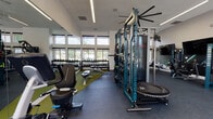 This contemporary fitness center is designed to support a comprehensive workout experience within a bright, open single-floor layout. The space is characterized by soaring windows along multiple walls, flooding the interior with abundant natural light and offering views of an outdoor pool area and surrounding greenery. Linear LED lighting runs across the ceiling, complementing the natural illumination throughout.

The cardio zone features a row of Pro Maxima treadmills positioned along the windowed wall, providing an energizing view during use. Nearby, an elliptical trainer and additional cardio equipment round out the aerobic offerings. A dramatic, large-scale photographic mural of a mountain trail runner dominates one wall, adding a dynamic and motivating visual focal point to the space.

The strength training area is anchored by a teal-accented functional trainer/cable machine system, a lat pulldown station, and a full rack of dumbbells arranged neatly along a mirrored wall. Adjustable weight benches, a trampoline, and medicine balls provide versatile training options. Rubberized flooring and green turf matting define distinct workout zones, enhancing both safety and functionality. A ceiling fan ensures comfortable air circulation. Double glass entry doors open to the exterior, reinforcing the space's connection to the outdoors.
