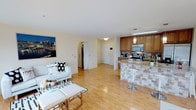 This single-floor space showcases an open-concept layout that connects the living area and kitchen in one cohesive, light-filled environment. Warm hardwood flooring runs throughout, lending a clean and inviting feel to the entire space.

The living area is comfortably proportioned, featuring neutral cream-colored walls that maximize the sense of brightness. Decorative wall art adds a pop of color and personality to the space.

The kitchen is well-appointed with medium-toned wood cabinetry, stainless steel appliances including a range and microwave, and a tiled backsplash that adds visual texture. A peninsula with a granite-style countertop and tiled base serves as both a functional workspace and a casual dining area, complete with modern bar stools. A pendant light fixture above the peninsula provides focused task lighting.

A hallway visible from the main living space leads to additional rooms, suggesting a practical and efficient floor plan. The entry door is clearly visible, contributing to an intuitive flow throughout the unit. The overall ambiance is clean, modern, and welcoming.