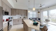 This contemporary single-floor residence showcases a thoughtfully designed open-concept layout that seamlessly connects the kitchen, dining, and living areas. The kitchen is outfitted with wood-tone cabinetry, a white subway tile backsplash, stainless steel appliances — including a full-size refrigerator, range, microwave, and dishwasher — and a large center island with a quartz countertop that doubles as a breakfast bar. Pendant lighting and track lighting illuminate the space, complementing the light-toned tile flooring found throughout. The living area benefits from soaring ceilings, large floor-to-ceiling sliding glass doors, and access to a private balcony overlooking a vibrant city skyline, flooding the interior with natural light. The primary bedroom features high ceilings, a neutral color palette with a bold accent wall, a walk-in closet, and a private en-suite bathroom with wood-tone vanity cabinetry. A second bedroom continues the modern aesthetic with a striking dark blue accent wall and contemporary finishes. A dedicated walk-in closet provides generous storage. The overall ambiance is clean, bright, and modern, with a cohesive neutral palette accented by contemporary fixtures and thoughtful design details throughout.