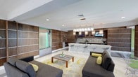 This single-floor amenity space showcases a sophisticated, contemporary design that seamlessly blends social and lounge functions. The layout opens with a welcoming lobby area featuring large floor-to-ceiling windows that flood the space with natural light, offering views of an outdoor pool area. Curved and modular seating arrangements, paired with patterned area rugs and curated wall art, create an inviting atmosphere throughout.

A standout water wall feature with green-tinted illumination anchors one side of the lounge, adding a tranquil, spa-like ambiance. Rich wood-paneled walls with horizontal detailing provide warmth against the otherwise cool, modern palette of whites and grays. Large-format tile flooring with varied grey tones runs throughout, unifying the space.

The wet bar area is a centerpiece of the social zone, featuring a sleek white quartz countertop, mosaic tile backsplash, and a row of clear acrylic barstools. Dramatic cluster pendant lights hang overhead, complementing mounted flat-screen televisions built into the dark wood cabinetry. The overall ambiance is polished and upscale, designed to encourage both relaxation and social interaction.