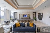 Located at 401 W 25th St in Houston's vibrant Heights neighborhood, this thoughtfully designed amenity floor offers a refined and welcoming environment for residents. The entry lobby greets visitors with large-format light stone tile flooring, recessed lighting, and a bold abstract artwork that sets a contemporary tone. A textured wood-panel accent wall and a reception counter anchor the space, while a tufted bench provides a functional yet elegant touch.

Moving into the main lounge, the design elevates with coffered ceilings featuring warm ambient uplighting and expansive floor-to-ceiling windows that flood the room with natural light and frame views of the surrounding neighborhood. A sleek marble-clad fireplace wall serves as the focal point, complete with a wall-mounted display above it. Plush seating arrangements, including a deep navy sofa, accent chairs, and a large tufted ottoman, create an inviting social atmosphere atop a richly patterned area rug.

Adjacent to the lounge, a bright café-style dining area features marble bistro tables paired with modern chairs, all bathed in sunlight from oversized windows overlooking open green space. A mosaic-tiled bar area and mirrored accent wall add stylish detail to this space. Completing the floor is a stainless steel elevator bank set against a striking stacked wood-look stone accent wall, lending an upscale, hotel-like quality throughout.