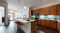 This single-floor residence offers a thoughtfully designed layout that balances functionality with modern aesthetic appeal. The living area is bright and welcoming, featuring dark hardwood-style flooring, a large window with transom detail above an exterior door, and views of the surrounding building exterior. The space receives ample natural light, creating an open and airy atmosphere.

The kitchen is a standout feature, showcasing warm-toned wood cabinetry extending to the ceiling, a green glass tile backsplash, and a full suite of stainless steel appliances including a range, microwave, and refrigerator. A generous island with a white quartz countertop provides bar seating and additional prep space, while pendant lighting and under-cabinet lighting add both function and ambiance. The kitchen opens directly to the living area, reinforcing the open-concept flow.

The bedrooms are carpeted and well-sized, each featuring large windows that bring in natural light. The primary bathroom includes large-format tile flooring, a dual-sink vanity with warm wood cabinetry and white countertops, a soaking tub, separate shower, and access to a walk-in closet. Throughout the residence, neutral tones and consistent finishes create a cohesive, contemporary living environment.