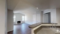 This single-floor residence offers a well-designed open-concept layout that seamlessly connects the main living areas. The spacious living room is a standout feature, highlighted by vaulted ceilings, a classic white-mantled fireplace, and multiple windows that invite abundant natural light. Sliding glass doors provide additional brightness and access to the outdoors. Dark hardwood-style flooring runs throughout the living and kitchen areas, adding warmth and a contemporary aesthetic.

The kitchen is thoughtfully equipped with white shaker-style cabinetry, dark granite countertops, recessed lighting, and a full suite of black appliances including a gas range, over-the-range microwave, and refrigerator. A curved granite-topped breakfast bar bridges the kitchen to the living space, making it ideal for casual dining or entertaining.

The bedroom features plush carpeting for added comfort, a textured ceiling, and direct sightlines to the kitchen. The bathroom is cleanly appointed with a granite vanity countertop, white cabinetry, and a large mirror. A dedicated laundry area is also visible within the floor plan. Throughout the residence, neutral gray walls and white trim create a cohesive, move-in-ready aesthetic.