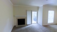 This single-floor space presents a clean, understated aesthetic with a neutral color palette throughout. The living area serves as the focal point of the space, highlighted by a classic white-mantled fireplace with tile surround and raised hearth, adding a traditional architectural element to the room. Crown molding lines the upper perimeter of the walls, contributing a refined detail to the otherwise simple interior.

Soft, neutral-toned carpeting extends across the living area, providing warmth underfoot and complementing the light cream walls. A textured ceiling adds subtle visual interest overhead. Natural light enters through a large sliding glass door fitted with vertical blinds, which opens directly onto a balcony enclosed by a metal railing. Views of surrounding greenery are visible through the door, offering a connection to the outdoors.

The overall ambiance of the space is calm and functional, with a traditional design sensibility. The combination of the fireplace, crown molding, and balcony access gives the space a comfortable, livable character suited for everyday use.