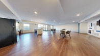 This single-floor community room offers a generous, open-concept layout that is well-suited for gathering, collaboration, and shared use. Rich hardwood flooring runs throughout the entire space, providing a warm and cohesive aesthetic. Recessed lighting and pendant fixtures illuminate the interior, complemented by ample natural light streaming in through multiple windows and a set of glass-paneled entry doors.

The kitchen area is equipped with dark cabinetry, a full-size refrigerator, a stove with range hood, and a light-colored countertop island with bar seating — ideal for food preparation and casual dining. Adjacent to the kitchen, a bright dining nook with glass doors leading outdoors provides an inviting spot for small group gatherings.

The main open area is arranged with several modular tables and modern upholstered seating, offering flexible configurations for meetings or social events. A wall-mounted flat-screen television is centered on a feature wall decorated with a geometric arrangement of blue, gray, and black acoustic panels, adding a contemporary and functional design element. A teal-framed double door provides secondary access, while clean white walls and a neutral color palette keep the overall atmosphere bright, professional, and welcoming.