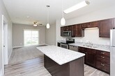 This single-floor residence offers a thoughtfully designed layout with modern finishes throughout. The kitchen serves as a standout feature, showcasing rich dark-espresso cabinetry that extends to the ceiling for ample storage, light granite countertops, and a full suite of stainless steel appliances including a range, microwave, dishwasher, and refrigerator. A central kitchen island with a matching granite surface provides additional prep space and doubles as a casual dining area, while pendant lighting adds a polished touch overhead. Light wood-style flooring runs throughout the kitchen and connecting areas, enhancing the bright and clean aesthetic. The bedroom is a comfortable, carpeted retreat featuring neutral taupe walls, a ceiling fan with integrated lighting, and a window that allows natural light to fill the room. The bathroom is well-equipped with a full soaking tub and shower combination with a curved curtain rod, a granite-topped vanity, and wood-style flooring that coordinates with the rest of the unit. A practical stacked washer and dryer unit is conveniently located within the bathroom, streamlining daily routines. Overall, the space presents a cohesive, contemporary feel with a neutral color palette and quality finishes.