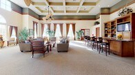 This expansive single-floor communal space exudes a warm, resort-inspired ambiance through its carefully considered architectural details and generous proportions. The centerpiece of the interior is a striking coffered ceiling with recessed lighting, finished in contrasting tones of cream and olive green, accented by rich wood crown molding that adds a refined, classic character throughout.

The main gathering area is carpeted with a neutral, patterned floor covering and furnished with comfortable lounge seating arranged to encourage conversation and relaxation. Tall, elegantly draped windows dressed in warm brown curtains line the walls, flooding the interior with abundant natural light and offering views of lush tropical landscaping and palm trees outside.

A built-in bar and entertainment area anchors one side of the room, featuring rich wood cabinetry with open shelving, a flat-screen television, and counter-height seating — ideal for social gatherings. A decorative ceiling fan adds a tropical, leisure-oriented aesthetic to the space.

The entryway is distinguished by a grand arched transom window, white columns, and tiled flooring, creating an impressive and welcoming arrival experience. Lush indoor tropical plants are thoughtfully placed throughout, reinforcing the vibrant, Florida-inspired atmosphere. Cozy seating nooks positioned beside the large windows provide inviting spots to take in the bright, garden-filled outdoor views.
