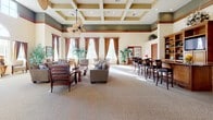 This expansive single-floor communal space exudes a warm, resort-inspired ambiance through its carefully considered architectural details and generous proportions. The centerpiece of the interior is a striking coffered ceiling with recessed lighting, finished in contrasting tones of cream and olive green, accented by rich wood crown molding that adds a refined, classic character throughout.

The main gathering area is carpeted with a neutral, patterned floor covering and furnished with comfortable lounge seating arranged to encourage conversation and relaxation. Tall, elegantly draped windows dressed in warm brown curtains line the walls, flooding the interior with abundant natural light and offering views of lush tropical landscaping and palm trees outside.

A built-in bar and entertainment area anchors one side of the room, featuring rich wood cabinetry with open shelving, a flat-screen television, and counter-height seating — ideal for social gatherings. A decorative ceiling fan adds a tropical, leisure-oriented aesthetic to the space.

The entryway is distinguished by a grand arched transom window, white columns, and tiled flooring, creating an impressive and welcoming arrival experience. Lush indoor tropical plants are thoughtfully placed throughout, reinforcing the vibrant, Florida-inspired atmosphere. Cozy seating nooks positioned beside the large windows provide inviting spots to take in the bright, garden-filled outdoor views.