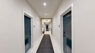 The common hallway at Milliner Lofts in Hatboro, Pennsylvania, makes a strong first impression with its thoughtfully designed interior that balances functionality with contemporary style. The space is finished with large-format white tile flooring, accented by dark runner rugs bearing the building's distinctive "M" monogram, guiding residents through the corridor with both practicality and flair.

The lobby hub anchors the space, featuring a stainless steel elevator set against a bold teal accent wall — a signature design element that carries throughout the hallway. A Milliner Lofts branded sign adds to the polished, branded atmosphere. Adjacent to the elevator, a sleek dark wood console provides a surface for packages and décor, while a built-in bank of individual mailboxes ensures organized mail service for all residents.

Ornate clustered light fixtures illuminate the entry area, adding an elegant, upscale touch to the otherwise clean and modern aesthetic. A set of gold-framed arched mirrors near the elevator enhances the sense of space. The residential corridor itself is lined with deep teal unit doors, recessed lighting, and crisp white walls, creating a clean and welcoming passage. Decorative vases and potted greenery are placed throughout, adding warmth and life to the overall environment.
