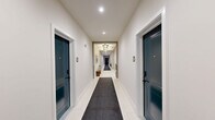 The common hallway at Milliner Lofts in Hatboro, Pennsylvania, makes a strong first impression with its thoughtfully designed interior that balances functionality with contemporary style. The space is finished with large-format white tile flooring, accented by dark runner rugs bearing the building's distinctive "M" monogram, guiding residents through the corridor with both practicality and flair.

The lobby hub anchors the space, featuring a stainless steel elevator set against a bold teal accent wall — a signature design element that carries throughout the hallway. A Milliner Lofts branded sign adds to the polished, branded atmosphere. Adjacent to the elevator, a sleek dark wood console provides a surface for packages and décor, while a built-in bank of individual mailboxes ensures organized mail service for all residents.

Ornate clustered light fixtures illuminate the entry area, adding an elegant, upscale touch to the otherwise clean and modern aesthetic. A set of gold-framed arched mirrors near the elevator enhances the sense of space. The residential corridor itself is lined with deep teal unit doors, recessed lighting, and crisp white walls, creating a clean and welcoming passage. Decorative vases and potted greenery are placed throughout, adding warmth and life to the overall environment.
