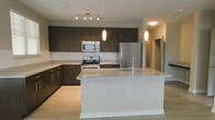 This single-floor space showcases a contemporary open-concept kitchen designed with both style and functionality in mind. The kitchen is anchored by a large central island topped with light granite countertops, complete with an integrated sink and a modern chrome faucet. Elegant pendant lights hang above the island, complementing the track lighting overhead to create a warm, well-lit environment.

Dark espresso-toned cabinetry lines the walls, providing generous storage while offering a striking contrast against the light granite countertops and white subway tile backsplash. The kitchen is equipped with a full suite of stainless steel appliances, including a gas range, over-the-range microwave, and a full-size refrigerator, catering to everyday culinary needs.

Light-toned hardwood flooring runs throughout, enhancing the bright and airy feel of the space. The open layout seamlessly connects the kitchen to the adjacent living areas, making the space ideal for entertaining. The neutral color palette and clean lines throughout contribute to a modern, polished aesthetic.