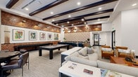 Located at 1300 Front Ave in Columbus, Georgia, this single-floor amenity lounge offers a generously sized, open-concept gathering space designed with a modern yet warm aesthetic. The interior is anchored by striking textured brick accent walls and a bold chevron-patterned feature wall that draws the eye toward the back of the room, where a built-in bar counter with navy blue bar stools provides a social hub. Exposed dark wood ceiling beams run throughout, complemented by a combination of recessed lighting, industrial-style pendant lights, and geometric diamond pendants that create a layered, inviting ambiance. The space is well-appointed for recreation and relaxation, featuring a full-size pool table, a shuffleboard table, and curated lounge seating areas with a mix of plush neutral sofas, leather armchairs, and warm amber accent chairs. A gallery wall of colorful framed artwork adds vibrancy along the brick-clad wall. On the opposite end of the space, floor-to-ceiling grid-pane windows flood a secondary lounge area with natural light and offer open views of the surrounding streetscape. A large-format dual flat-screen display mounted on a light stone-tile wall completes the entertainment offerings. The overall atmosphere is polished, welcoming, and purpose-built for community engagement.