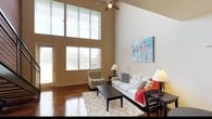 This two-floor residence is defined by its dramatic double-height living room, where soaring ceilings draw the eye upward and a generous bank of tall windows floods the space with natural light. A ceiling fan anchors the room, while polished stained concrete floors lend an industrial-modern character throughout the main level. The living area is open and inviting, offering ample room for comfortable seating arrangements beneath the expansive walls.

The kitchen is thoughtfully appointed with rich dark cabinetry, granite-look countertops, a double stainless steel sink, and a full suite of stainless steel appliances including a range, microwave, dishwasher, and refrigerator. The same stained concrete flooring continues here, creating a cohesive aesthetic.

A half bath on the main level features a standout exposed brick accent wall, adding warmth and texture, with a stacked washer and dryer conveniently located nearby.

Upstairs, the bedrooms are carpeted for comfort and each equipped with ceiling fans. The primary bedroom is notably spacious, featuring an exposed brick accent wall, a sliding window with outdoor views, and an open loft-style overlook. A second bedroom offers a cozy, angled ceiling detail. A full bathroom on the upper level includes a tub-shower combo, tile flooring, and dark vanity cabinetry with granite-look countertops.