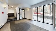 Located at 500 Avenue des Pins Ouest in Montréal's vibrant Mile End–Plateau neighbourhood, this building lobby serves as a functional and welcoming entrance space. The single-floor layout is organized into distinct zones that guide residents and visitors through the entry sequence efficiently.

The vestibule greets arrivals through a set of large glass doors framed in sleek aluminum, providing a clear visual connection to the street. Dark large-format tile flooring and carpet runners define the transition zones, while white walls and recessed lighting maintain a clean, bright atmosphere throughout.

Moving inward, the mailbox alcove features an extensive bank of individual metal mailboxes mounted on white walls, complemented by transom windows that introduce natural light from above. Baseboard heaters line the walls, a practical feature well-suited to Montréal's climate.

A raised carpeted section with dark tile risers leads further into the lobby, where an elegant arched ceiling detail adds architectural character. A large ornate black-framed mirror enhances the sense of depth in this area, while a full-height window offers a direct view to the exterior streetscape.

Dark wainscoting tiles line the corridor walls, creating a contrast against the crisp white upper walls and ceilings. Recessed and flush-mount ceiling fixtures ensure consistent, well-distributed illumination. The overall ambiance is functional yet refined, reflecting a well-maintained residential building entrance in one of Montréal's most sought-after neighbourhoods.