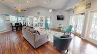 This single-story clubhouse is designed to serve as a functional and inviting community hub. Rich hardwood flooring flows throughout the entire space, complemented by soft gray walls and soaring vaulted ceilings that lend an open, airy feel to every area.

The main lounge is a generously proportioned gathering space furnished with a plush gray sofa and vibrant teal accent chairs arranged around a patterned area rug. A wall-mounted flat-screen TV, pendant lighting, and a ceiling fan add to the room's comfort, while abundant natural light pours in through multiple windows and glass-paned French doors.

Adjacent to the lounge, a dining area features a round glass-top table with dark wood chairs and is flanked by windows overlooking the outdoors. Informational displays line the walls, making this a practical spot for community announcements.

The fitness center, accessible through French doors, is equipped with cardio machines including a treadmill and is brightened by large windows. The leasing office area includes a full workstation with seating for visitors, surrounded by natural light from surrounding windows.

A kitchenette rounds out the amenities, boasting white cabinetry, a mosaic tile backsplash, a breakfast bar with teal barstools, and direct access to an outdoor patio via French doors. Together, these spaces create a cohesive and well-appointed clubhouse experience.