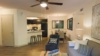This single-story residence showcases an open-concept floor plan that integrates the living, dining, and kitchen areas into one cohesive, functional space. Hardwood-style flooring extends throughout, providing a warm and consistent aesthetic across all zones.

The kitchen is appointed with dark cabinetry, a tile backsplash, granite-style countertops, and black appliances, including a refrigerator and range. A breakfast bar with bar stool seating offers a casual dining option and serves as a natural divider between the kitchen and the living area.

The dining area accommodates a full table and seating, illuminated by a contemporary pendant light fixture. Adjacent to it, the living area provides comfortable seating space, enhanced by a ceiling fan with an integrated light kit that contributes to both airflow and ambient lighting.

The neutral wall tones serve as a versatile backdrop throughout the unit, complementing the darker accents of the cabinetry and furnishings. The open layout promotes a sense of spaciousness, making the overall environment feel bright, connected, and well-suited for both everyday living and entertaining.