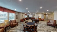 This well-appointed community activity room is designed to accommodate a variety of social and recreational pursuits within a single, open floor plan. The space is organized into several distinct functional zones that flow seamlessly into one another.

A dedicated screening area anchors one end of the room, featuring a ceiling-mounted projector and a large pull-down screen, flanked by rows of plush, upholstered armchairs and sofas arranged in a theater-style configuration. Warm-toned curtain valances frame the windows, admitting natural light while maintaining a cozy atmosphere.

Adjacent to the screening area, two octagonal game tables with leather-padded chairs provide space for cards or board games. A full-length shuffleboard table occupies its own alcove, accented by a gallery-style row of framed artwork along the wall above it.

Multiple lounge seating arrangements are distributed throughout the room, including accent chairs in earthy terracotta and tan tones paired with decorative side cabinets. A wall-mounted flat-screen television creates an additional viewing area within one of these lounge clusters.

Recessed ceiling lighting illuminates the entire space evenly, while patterned, neutral-toned carpeting ties all zones together. Decorative wall accents, potted plants, and framed artwork contribute to the room's polished, welcoming ambiance.