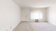 Located at 2970 East Ruby Drive in Fullerton, CA, this single-story, one-bedroom, one-bathroom unit offers a functional and efficient floor plan. The living room is carpeted and bathed in natural light from a large window dressed with vertical blinds, with a wall-unit air conditioner providing climate control. A hallway connects the main living areas, transitioning from carpet to hardwood-style flooring as you move toward the kitchen and dining area. The combined dining room and kitchen features a galley-style kitchen equipped with white cabinetry, dark granite-style countertops, a gas range, and a single-basin sink — all set on light hardwood-style flooring. An adjacent built-in cabinetry unit with glass-front upper cabinets and a ceiling fan adds charm and storage to the dining area. The spacious primary bedroom is fully carpeted and features a large window with vertical blinds and a wall-unit for temperature control. The bathroom includes a wide vanity with ample counter space, white cabinetry, a large mirror, and hardwood-style flooring. A dedicated closet provides additional storage. The overall aesthetic is clean and bright, with white walls and neutral tones throughout.