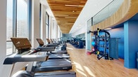 This well-appointed fitness center in Bethesda, MD offers a generously sized, single-floor workout space designed with both form and function in mind. The facility is immediately distinguished by its soaring ceilings adorned with elegant linear wood-slat paneling, which lends warmth and a modern aesthetic to the environment. Floor-to-ceiling windows line the perimeter, flooding the space with natural light and providing open views of the surrounding Bethesda landscape.

The layout is thoughtfully organized into distinct workout zones. A long row of treadmills, ellipticals, and cardio machines runs along the windowed wall, allowing users to exercise while enjoying the outdoor views. The strength training area features a comprehensive selection of selectorized weight machines with tan upholstered seating, free weights including a full dumbbell rack, and adjustable benches. Functional training equipment — including resistance bands, medicine balls, and cable machines — is neatly organized along dedicated racks.

The flooring transitions between warm hardwood and rubber sport tile, delineating cardio and strength zones respectively. Large wall mirrors expand the sense of space and support proper form during workouts. The overall ambiance is bright, clean, and professional, making this fitness center a premier destination for health and wellness in Bethesda.