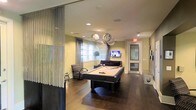 This single-floor recreational lounge is designed for both entertainment and relaxation, combining modern aesthetics with functional amenities. The centerpiece of the space is a full-sized pool table finished in dark wood, positioned beneath a cluster of decorative globe pendant lights that serve as a striking focal point. The warm hardwood flooring extends throughout, grounding the room with a rich, polished look.

The lounge area is furnished with plush seating, including armchairs and a sofa, arranged near a wall-mounted flat-screen television, creating a comfortable viewing nook. Large windows along the far wall allow natural light to brighten the interior, complementing the soft yellow-toned walls that contribute to a welcoming ambiance.

A sheer, metallic chain curtain partition near the entrance adds a decorative element that subtly defines the boundary of the space while maintaining an open feel. Recessed ceiling lighting supplements the pendant fixtures, ensuring the room is well-lit throughout. A glass-paneled door at the rear provides access to an adjacent area, enhancing the space's connectivity and functionality.