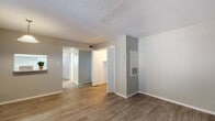 This single-floor unit presents a straightforward, functional layout with a cohesive neutral palette throughout. The main living area is finished with wood-look plank flooring in a warm tone, complemented by light gray walls and clean white trim. A textured ceiling adds subtle detail overhead, while a wall-mounted HVAC unit and visible vent registers speak to the space's practical utility. Open doorways connect the living area to a hallway, leading to the private rooms beyond.

A secondary living or flex space features similar wood-look flooring in a slightly deeper tone, offering a versatile area that can serve multiple purposes. The bedroom is carpeted in a neutral gray tone and includes a ceiling fan with integrated lighting, as well as a window fitted with blinds that allows natural light to filter into the room.

The bathroom is finished in a clean, all-white scheme with wood-look flooring, a single-sink vanity topped with a large mirror, a well-lit vanity light bar, and a full bathtub with a tiled surround. The overall design is simple and well-maintained, offering a move-in-ready environment with consistent finishes across each room.