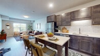 This single-floor unit is designed with a contemporary aesthetic and a functional open-concept layout. The kitchen serves as a central feature, equipped with dark espresso-toned shaker-style cabinetry reaching toward the ceiling, white subway tile backsplash, light quartz countertops, and a full suite of stainless steel appliances including a refrigerator, dishwasher, microwave, and range. A freestanding kitchen island with seating provides additional prep space and casual dining.

The kitchen flows directly into the living area, which features wood-style plank flooring, neutral walls, and a window offering natural light and outdoor views. The bedroom spaces are carpeted and include windows that bring in additional daylight. One bedroom benefits from a generous walk-in closet outfitted with multi-tier wire shelving and hanging rods across several walls.

The bathroom is well-sized, featuring a dark vanity with a white countertop, a wide mirror, wood-style flooring, and a full tub-shower combination. Throughout the unit, the consistent use of dark wood tones against light finishes creates a cohesive, modern contrast that gives the space a polished and inviting character.