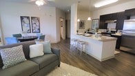 This single-floor residence showcases an open-concept layout that seamlessly connects the living and kitchen areas, creating a cohesive and functional living environment. The living area features rich, dark hardwood-style flooring that extends throughout the space, anchored by a plush area rug and a comfortable sofa accented with decorative throw pillows. A ceiling fan overhead provides both comfort and light, while framed wall art adds a polished, curated feel to the space.

The kitchen is positioned along one side of the open floor plan and is highlighted by a peninsula-style breakfast bar with light countertops, accommodating three modern metal barstools. Pendant lights hang above the bar, adding a contemporary touch and warm illumination. Dark cabinetry contrasts attractively with the light countertops, and modern appliances complete the functional kitchen setup.

A dining nook with a small table and chairs sits adjacent to the living area, making use of the open layout efficiently. A hallway visible in the background leads to additional rooms, suggesting a well-organized floor plan. The neutral wall tones and consistent flooring throughout lend the space a bright, airy, and cohesive aesthetic.