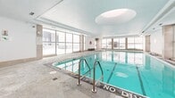 This indoor aquatic facility offers a well-appointed swimming environment spanning a substantial footprint, designed to accommodate both recreational and lap swimming. The centerpiece is a clearly marked lap pool with designated swim lanes, stainless steel entry railings, and clearly posted depth and safety signage, including "No Diving" and "Caution No Diving" notices throughout.

The space is bathed in natural light courtesy of large floor-to-ceiling windows that line multiple walls, providing an open, airy atmosphere. Oval skylights in the paneled ceiling further enhance the brightness of the interior, creating a clean and refreshing ambiance. The ceiling itself features a tiered, structured design with integrated ventilation, contributing to the facility's polished, modern aesthetic.

The surrounding pool deck is finished in a textured, slip-resistant material, providing ample space for movement around all sides of the pool. Poolside amenities include benches for seating, a life ring station, and a staffed or supervised counter area visible near the entrance. Safety equipment is mounted prominently on the walls for easy access.

The overall design is clean and functional, with neutral tones of white, light grey, and warm beige accents framing the vivid turquoise water of the pool. The facility presents a well-maintained, professional-grade aquatic environment suitable for regular swimming and fitness use.