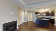 This well-appointed single-floor residence offers a thoughtfully designed open-concept layout that seamlessly connects the living and kitchen areas. The living space is anchored by a sleek fireplace set against neutral gray walls, creating a warm and inviting atmosphere. Rich, dark hardwood flooring runs throughout, adding a sense of warmth and continuity to the space.

The kitchen is a standout feature, equipped with stainless steel appliances including a refrigerator, stove, and microwave. Dark espresso cabinetry paired with a light granite countertop island creates a striking contrast, while stylish pendant lighting suspended above the island adds both function and ambiance. The kitchen's efficient design maximizes workspace and storage.

A tray ceiling detail in the living area adds architectural interest and a touch of elegance, enhancing the sense of height and openness. Recessed lighting complements the pendant fixtures, ensuring the space is well-lit throughout. Doorways leading to additional rooms suggest a practical and well-organized floor plan. The neutral color palette of warm beige and cool gray tones throughout creates a versatile, move-in-ready environment suitable for a variety of interior styles.
