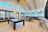 Located in Aurora, CO, this contemporary community amenity space is designed to impress and accommodate. The room is anchored by dramatic vaulted ceilings with exposed natural wood beams, soaring to considerable heights and amplifying the open, airy feel of the space. Light hardwood flooring runs throughout the main area, complementing the crisp white walls and modern aesthetic.

A centerpiece pool table sits prominently in the main gathering area, positioned beneath a striking starburst chandelier that serves as a bold decorative focal point. An abundance of natural light pours in through multiple large windows and clerestory windows positioned high along the walls, offering views of the surrounding Aurora neighborhood.

The lounge and dining area features a mix of seating arrangements, including a built-in blue upholstered banquette, wooden communal tables, and contemporary chairs — ideal for casual dining or social gatherings. Sleek pendant lights hang above, adding warmth to the space.

A dedicated kitchenette area is finished with light wood cabinetry, white quartz countertops, and a bold dark glazed tile accent wall with wall sconces. A built-in beverage station and under-counter refrigerator complete this functional nook. The overall design blends sophistication with comfort, creating an inviting, multi-purpose environment suited for community interaction and recreation.