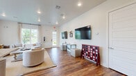 Located in Crandall, TX, this single-floor space offers an open-concept layout that seamlessly integrates the kitchen and living areas. The living room is bathed in natural light, courtesy of large multi-pane windows dressed with flowing neutral curtains that frame views of an outdoor pool and lush greenery. Warm hardwood flooring runs throughout the space, anchoring the clean, neutral palette of the walls and trim.

The living area is welcoming and versatile, featuring recessed lighting, a woven area rug, and ample room for comfortable seating arrangements. A dedicated media wall with mounted flat-screen televisions offers a functional entertainment setup, while a side entrance door provides convenient outdoor access.

The kitchen is well-appointed with white and grey shaker-style cabinetry, granite countertops, and a full suite of stainless steel appliances including a refrigerator, dishwasher, and microwave. A peninsula with granite countertop serves as a natural divider between the kitchen and living area and is accented by a stylish gold pendant light. Recessed lighting further brightens the space, making it both practical and inviting. Together, these spaces create a cohesive, modern environment suited for everyday living and entertaining.